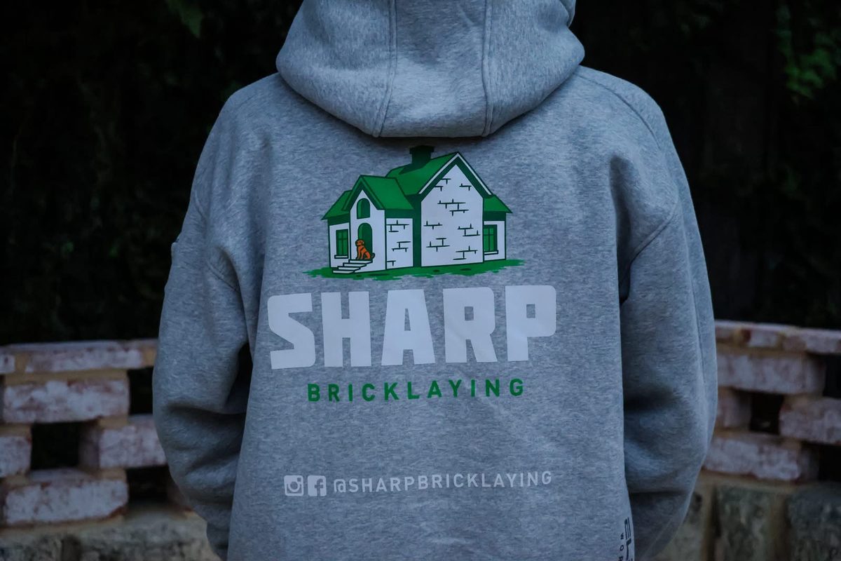 Back of a grey Sharp Bricklaying hoodie with the green house logo, in front of a breeze block wall